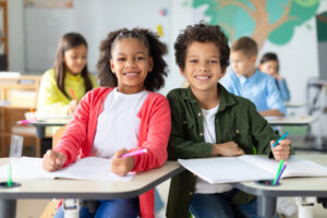 duas crianças sentadas em sala de aula sorrindo para a câmera. estão com seus cadernos abertos sobre a mesa
