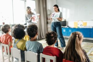 Duas professoras de pé ensinando a cinco alunos que estão sentados lado a lado em cadeiras