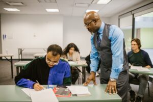 Um professor olhando para o aluno que está escrevendo em seu caderno [o professor está em pé no corredor]