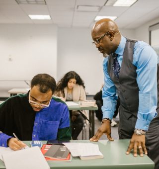 Um professor olhando para o aluno que está escrevendo em seu caderno [o professor está em pé no corredor]