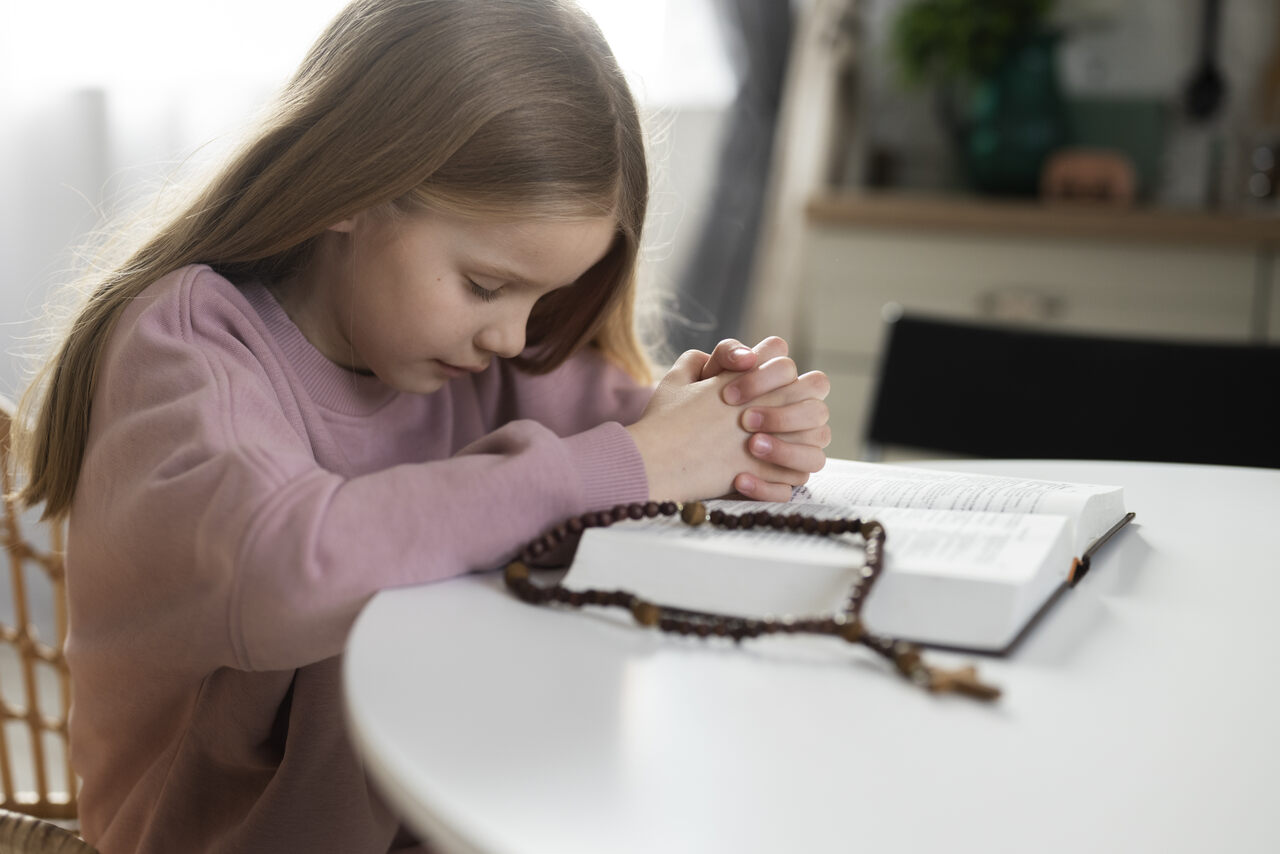 Menina orando com bíblia aberta na mesa e um terço em cima