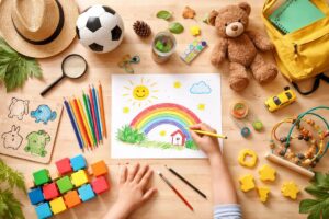 Criança desenhando arco-íris com lápis de cor sobre mesa de madeira cercada por brinquedos educativos, bola de futebol, ursinho de pelúcia, mochila amarela e materiais escolares, representando atividades para criança de 7 anos voltadas ao desenvolvimento criativo e coordenação motora