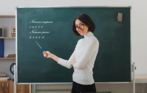 sorridente jovem professora de oculos em pe na frente do quadro negro e aponta para o quadro negro com ponteiro em sala de aula e1693339285411