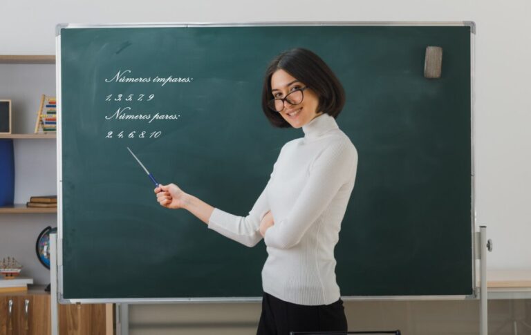sorridente jovem professora de oculos em pe na frente do quadro negro e aponta para o quadro negro com ponteiro em sala de aula e1693339285411