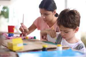 Atividades para Criança de 2 e 3 Anos (Aprender Brincando) Duas crianças na escola brincando. Elas estão sentadas e usam materiais para fazer pinturas