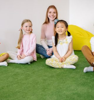 Uma professora e seus alunos sentados dentro de uma sala de aula em cima de um tapete verde