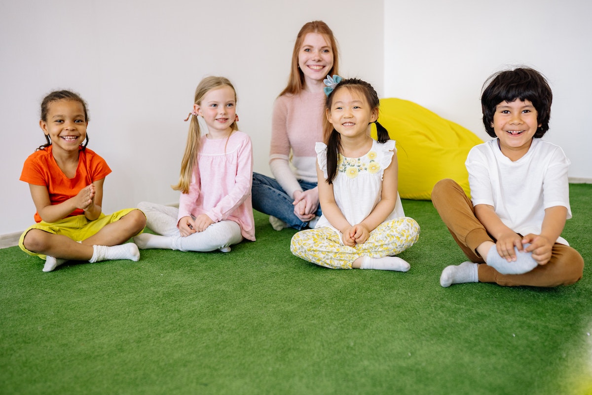 Uma professora e seus alunos sentados dentro de uma sala de aula em cima de um tapete verde
