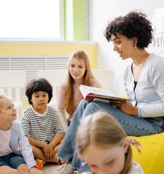 Uma professora sentada em um puff lendo uma história para seus alunos, que são crianças