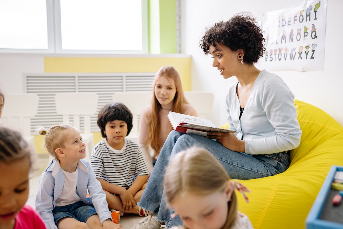 Uma professora sentada em um puff lendo uma história para seus alunos, que são crianças