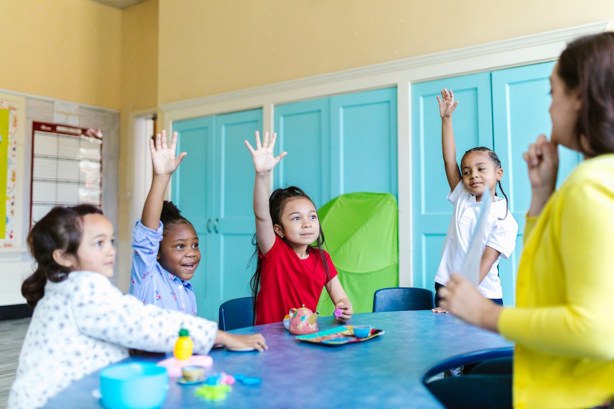Quatro alunos em uma sala de aula com as mãos levantadas como se quisessem fazer uma pergunta