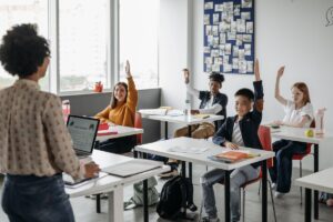 Atividades Sobre o Dia do Trabalho 2024: PDF para Imprimir Uma professora na frente da sala de aula com uma série de alunos com as mãos levantadas