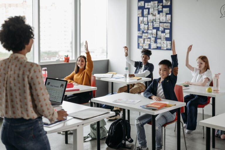 Uma professora na frente da sala de aula com uma série de alunos com as mãos levantadas