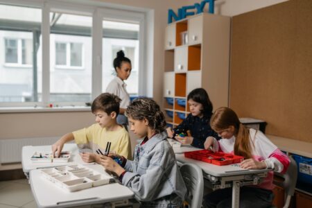 Quatro alunos sentados em uma sala de aula, e a professora está em pé ao fundo