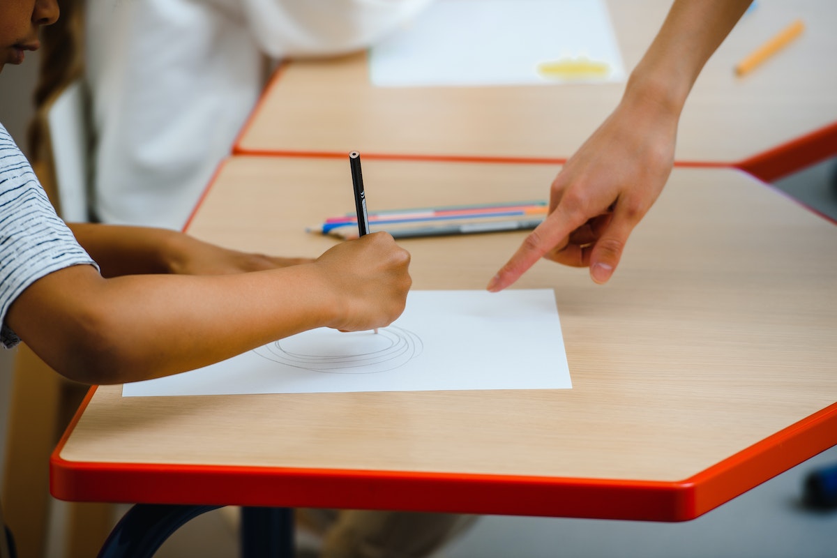 O dedo de uma professor aparece apontando na mesa para orientar o aluno que está pintando