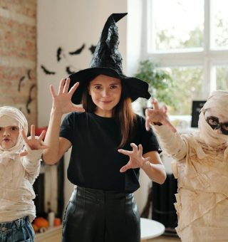 Três crianças estão fantasiadas. A menina no meio está de bruxa, e duas crianças menores estão de múmia