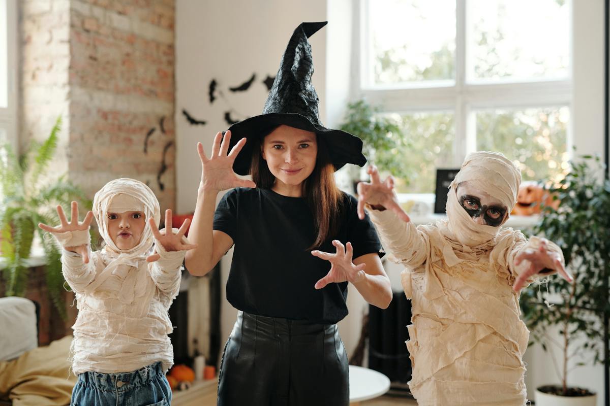 Três crianças estão fantasiadas. A menina no meio está de bruxa, e duas crianças menores estão de múmia