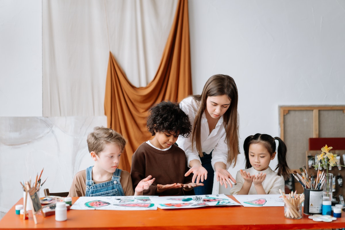 Uma professora está atrás de três crianças que estão sentadas pintando. Ela orienta os alunos com as cores