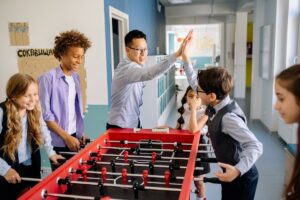 Alguns alunos estão jogando pebolim. Um professor comemora, tocando na mão do aluno que fez gol