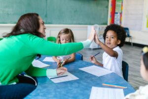Uma professora está tocando na mão de um de seus alunos, no fundo há outra aluna pintando