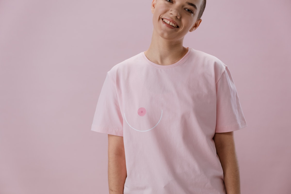 Uma mulher que está sorrindo. Ela está com o cabelo raspado e veste uma camisa rosa com um apoio à atividades sobre Outubro Rosa 2024