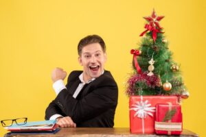 Brincadeiras de Natal para Fazer na Escola 2024 (Fundamental) Um professor está sentado e comemorando. Ao lado há uma árvore e alguns enfeites de Natal