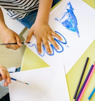 Uma criança está pintando em uma folha branca. Ela faz o contorno da sua mão com uma tinta azul