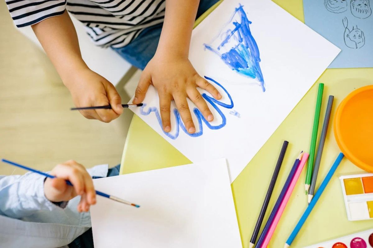 Uma criança está pintando em uma folha branca. Ela faz o contorno da sua mão com uma tinta azul