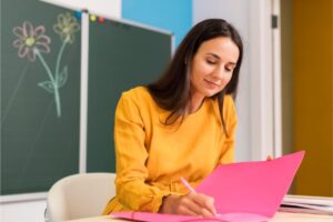 Uma professora está sentada em sua mesa enquanto faz anotações em um caderno