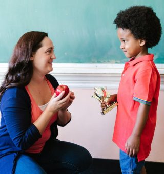 um aluno está entregando um presente para a professora, que está agachada e segura uma maçã nas mãos