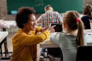 Uma professora agachada ao lado do aluno na sala de aula explicando a matéria