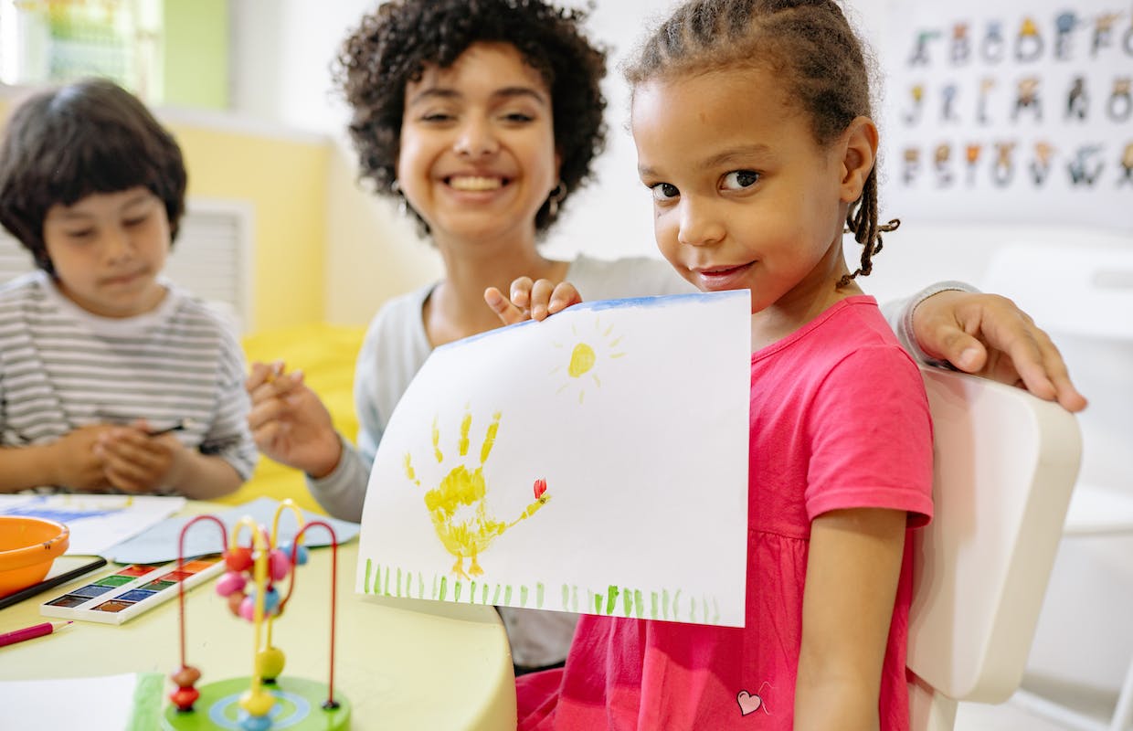 Uma professora e sua aluna estão na aula de pintura, a aluna segura uma folha em suas mãos onde tem um desenho feito com tinra