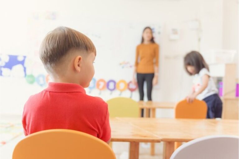 Imagem de costas de um aluno sentado na sala de aula. Ao fundo é possível ver a professora em pé e uma aluno também