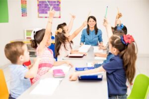Uma professora está sentada na mesa. Ao redor dela, diversos alunos estão com as mãos levantadas