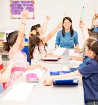 Uma professora está sentada na mesa. Ao redor dela, diversos alunos estão com as mãos levantadas