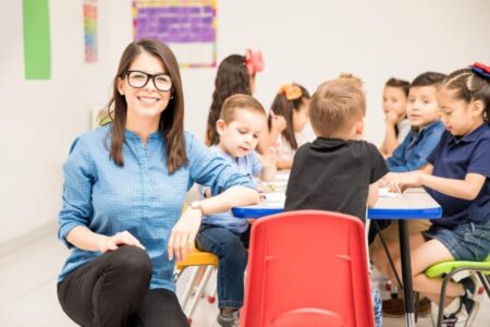 Atividade Letra J para Educação Infantil: Modelos Didáticos Uma professora sentada sorri para câmera enquanto está de costas para a turma que aparece ao fundo desfocada