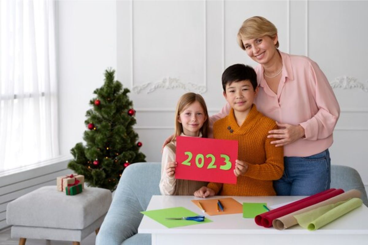 Uma mãe com seus dois filhos. Os filhos seguram um cartão de natal onde está escrito "2023" em uma folha A4