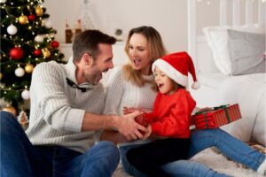 Uma família reunida comemorando o Natal. A criança está feliz no colo da mãe, e o pai entrega um presente para a garota