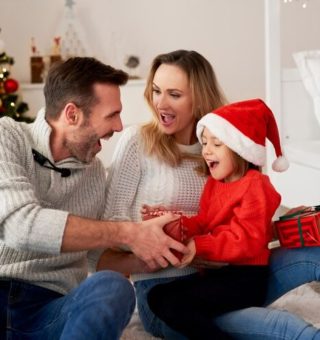 Uma família reunida comemorando o Natal. A criança está feliz no colo da mãe, e o pai entrega um presente para a garota