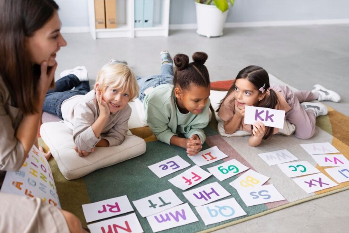 Três alunas deitadas na sala de aula. Uma delas segura uma placa de papel com a letra "kk"