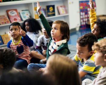 Uma turma de crianças sentadas na sala de aula. Algumas delas levantam a mão para fazer pergunta