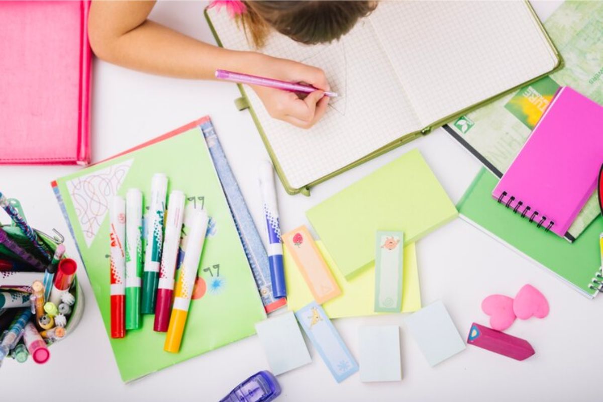 Uma aluna vista de cima escrevendo em um caderno. Na mesa dela há muitos materiais que ela usa para montar seu Portfólio Educação Infantil 2024