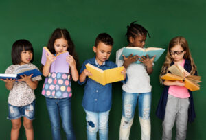Datas comemorativas de agosto: principais eventos para atividades Crianças em pé olhando os livros abertos que tem em suas mãos. Ao fundo uma parede verde escura simbolizando o quadro escolar