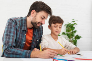 momento pai e filho fazendo atividades escolares juntos. Estão olhando para os cadernos com lápis nas mãos, o pai está ensinando algo ao menino