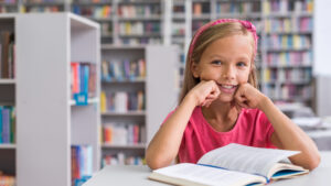 menina sentada em uma biblioteca com livro aberto sobre a mesa. Ela está sorrindo para a câmera com as mãos abaixo do queixo. Ao fundo é possível identificar estantes de livros coloridos
