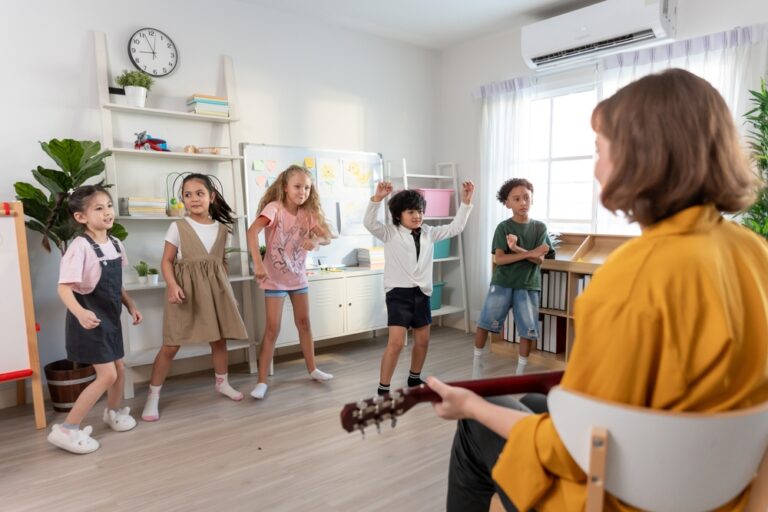 Crianças em sala de aula em pé, dançando enquanto uma professora toca violao