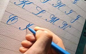 Mão segurando uma caneta azul enquanto escreve letras cursivas em um caderno pautado, representando atividades de caligrafia criativa e artística com foco em treino de escrita manual.